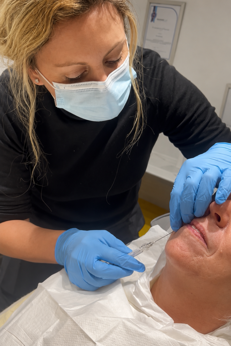 Healthcare worker in a mask and blue gloves administers an injection to a patient lying down; syringe near the patient's face.